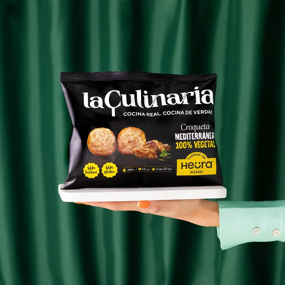 Mano que sostiene una bolsa de croquetas La Culinaria ante un telón verde.