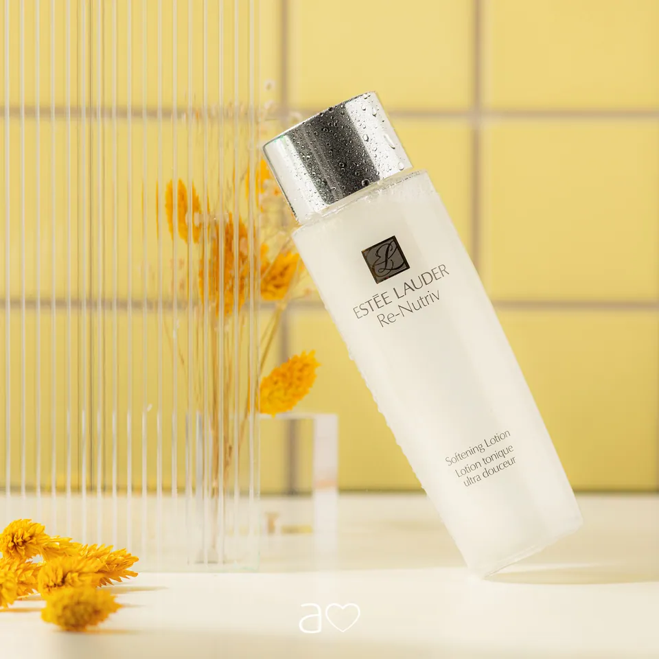 Frasco blanco de Estée Lauder ante vidrio acanalado y flores amarillas.